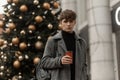 Urban hipster man in an elegant plaid coat with a stylish hairstyle stands and holds in hand a cup with a hot tea near the festive