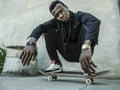 Young attractive and serious black African American man squatting on skate board at grunge street corner looking cool posing in Royalty Free Stock Photo