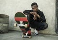 Urban portrait of young attractive and serious black afro American man with skate board squatting on street corner looking cool Royalty Free Stock Photo