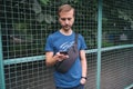 Urban lifestyle. Young bearded man in blue t-shirt with crossbody bag staying near fence of city sport playground and using