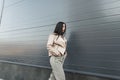 Urban pretty young woman in stylish oversized leather beige jacket in fashionable jeans walks near gray building in the city. Royalty Free Stock Photo