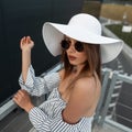 Urban stylish hipster woman in a stylish summer hat with round sunglasses in a fashionable striped top stands on the street in the Royalty Free Stock Photo