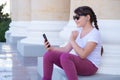 Urban stylish teenager girl sits near column, makes video call on mobile phones, uses hands free, talks with friends.