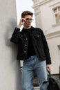 Urban stylish young hipster man in denim black jacket in jeans with leather backpack stands near vintage wall and straightens