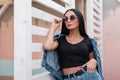 Urban stylish young woman hipster in a trendy black top in a fashionable denim jacket in a skirt in dark sunglasses posing