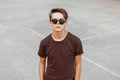 Urban trendy young hipster man in stylish brown t-shirt with hairstyle in trendy black sunglasses stands at the stadium. Royalty Free Stock Photo