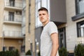 Urban young handsome man fashion model in a fashionable summer t-shirt stands on the street near a modern building. Nice guy walks