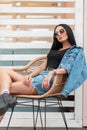 Urban young hipster woman in black leather boots in fashionable denim clothes in stylish sunglasses poses on a straw chair Royalty Free Stock Photo