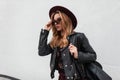 Urban young hipster woman in trendy sunglasses in a chic hat in a leather jacket with a black stylish backpack Royalty Free Stock Photo