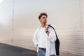 Urban young man fashion model with trendy hairstyle in a vintage white shirt in jeans with a black fabric bag relaxes on the Royalty Free Stock Photo