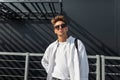 Urban young man hipster in trendy white shirt in fashionable sunglasses with a cloth black bag posing on a sunny day near a gray Royalty Free Stock Photo