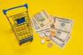 US dollars and cents next to the supermarket shopping trolley. Yellow background. Concept of shopping, discounts, online shopping