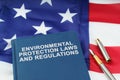 On the US flag lies a pen and a book with the inscription - ENVIRONMENTAL PROTECTION LAWS AND REGULATIONS