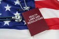 On the US flag lies a stethoscope and a book with the inscription - HEALTH LAWS AND REGULATIONS