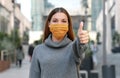 Use the mask! Young woman wearing protective face mask showing thumbs up with modern city street on the background