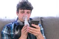 Use nebulizer and inhaler for the treatment. Sick man inhaling through inhaler mask and looking at mobile phone sitting