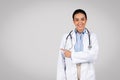 Useful information. Portrait of cheerful latin female doctor pointing at copy space, grey background, smiling at camera