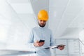 Using phone. Engineer in white clothes and orange protective hard hat standing and working indoors