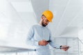 Using phone. Engineer in white clothes and orange protective hard hat standing and working indoors