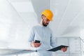 Using phone. Engineer in white clothes and orange protective hard hat standing and working indoors