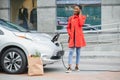 Using smartphone while waiting. African American girl on the electric cars charge station at daytime. Brand new vehicle