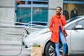 Using smartphone while waiting. African American girl on the electric cars charge station at daytime. Brand new vehicle