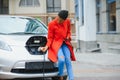 Using smartphone while waiting. African American girl on the electric cars charge station at daytime. Brand new vehicle