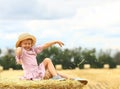 ÃÂ¡ute girl smiles in a wicker hat and a pink blue dress throws hay up sitting on a huge haystack. Childhood. Rest in the village.