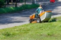 A gardener using a professional lawn mower mows tall green grass along of a city road