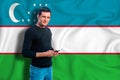 Uzbekistan flag on the background of the texture. The young man smiles and holds a smartphone in his hand. The concept of design