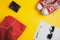 Vacation equipment. Top view of red shorts, leather wallet, denim jacket, sunglasses and notebook with pen on yellow background