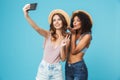 Vacation photo of two multiethnic girls wearing straw hats smiling and showing peace sign at camera, while taking selfie on smart Royalty Free Stock Photo