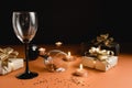 Two clinking wine glasses, various handmade gift boxes decorated with confetti, candle on orange and black background.