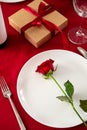 Valentine`s Day dinner table setting. Rose on white plate, gift box, cutlery, wine, glasses on red background Royalty Free Stock Photo