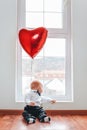 Valentine`s day. Little baby boy looks out the window, with a red ball in the form of a heart. Vertical Royalty Free Stock Photo