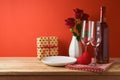 Valentine`s day romantic dinner concept. Wooden table with plate, wine glasses, gift box and rose flowers over red background Royalty Free Stock Photo