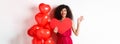 Valentines day and love concept. Cheerful young woman in elegant red dress, standing near romantic balloons and holding Royalty Free Stock Photo