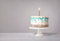 Vanilla Birthday Cake with a lit gold candle, teal blue drip and colorful sprinkles on a light grey white background Royalty Free Stock Photo