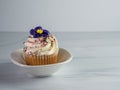 Vanilla cupcake with swirled white frosting, a purple fondant flower and sprinkles sitting in a white ceramic dish on a sandstone Royalty Free Stock Photo