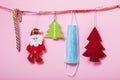 Various Christmas tree toys and a medical mask hang on a decorative ribbon on a pink background