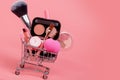 Various make-up products and brushes in shopping cart on pink background. Makeup cosmetics sale concept Royalty Free Stock Photo