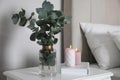 Vase with beautiful eucalyptus branches, book and candles on nightstand in bedroom