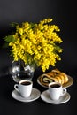 Vase with mimosa, two cups of coffee and poppy seed strudel on a black background