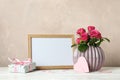 Vase with pink roses, empty frame, gift and little hearts on white table