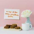Vase with white rose, tea cup, biscuits in front of pale pastel pink background. Card with inspirational quote