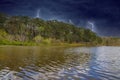 A vast rippling lake surrounded by lush green trees and bare winter trees on the banks with powerful storm clouds and lightning