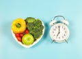 Vegetables capsicum, broccoli, tomatoes and apple in heart shape plate on blue background with white vintage alarm clock,