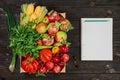Vegetables and fruits and herbs in a box layout on a dark wooden table, next to a notebook and a pencil for keeping track of the