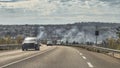 Vehicles driving on a fast lane, in the back the city from which smoke escapes