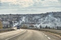 Vehicles driving on a fast lane, in the back the city from which smoke escapes